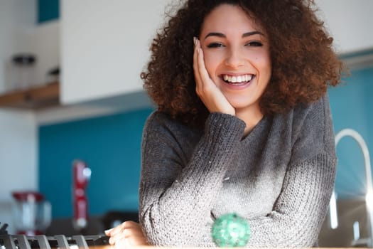 Smiling woman with curly hair leans on hand in a brightly lit kitchen. A blurred Christmas ornament in the foreground adds a festive touch. Her expression radiates warmth and joy
