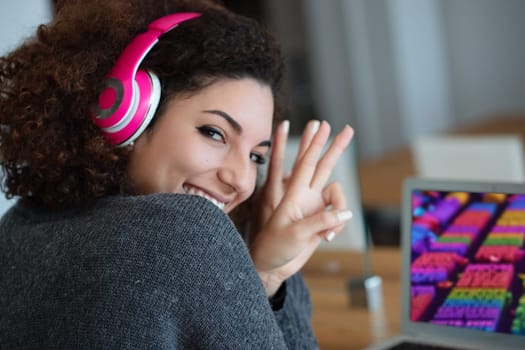 Young woman with curly hair happily listens to music on headphones in a modern office, flashing an approving ok sign while fully immersed in her work