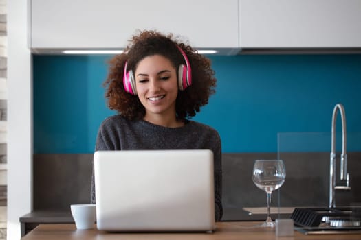 Young woman happily works on her laptop in a modern kitchen, typing, listening to music, and enjoying coffee. The cozy atmosphere and stylish design make it perfect for remote work
