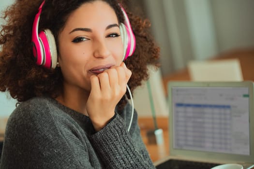 Young woman with curly hair is sitting at a table, listening to music and biting her nails while working on a laptop computer