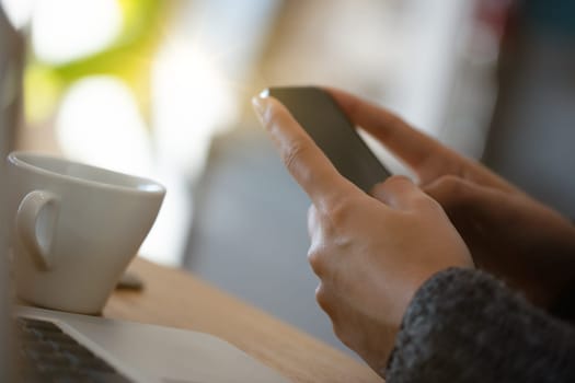 Woman is holding a mobile phone in her hands while sitting at a kitchen counter with a laptop and a cup of coffee nearby
