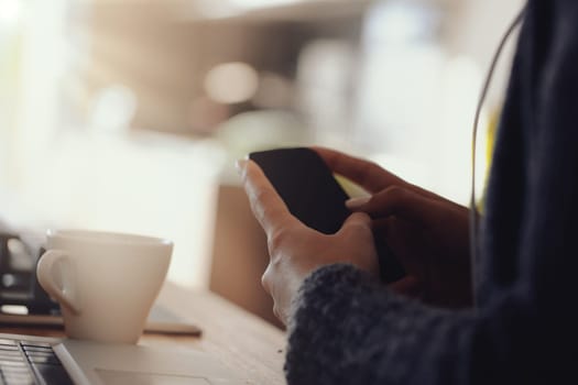 Hands hold a smartphone with a blank screen in a well-lit room. Nearby, a white coffee cup and other objects on a wooden table suggest a calm, casual setting with modern technology