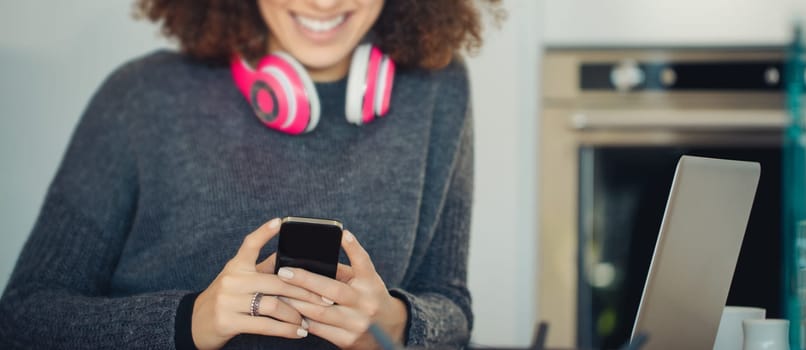 Young woman happily smiles in her kitchen, using a smartphone and wearing headphones, connecting through technology and social media with positive energy
