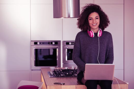 Young woman with curly hair sitting on her modern kitchen counter using a laptop and wearing headphones, working or studying from home