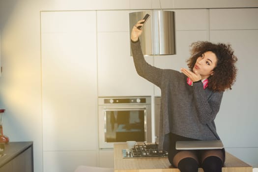 Young woman with curly hair takes a selfie in a bright kitchen, exuding a cheerful vibe. She sends a kiss to the camera, fully immersed in the digital world