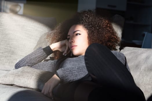 Curly-haired woman lounges on her couch in the living room, lost in thought. Her introspective expression indicates she's reflecting on profound and wonderful ideas, enjoying a moment of quiet contemp