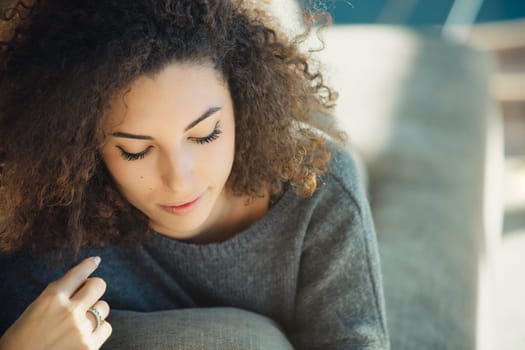 Young woman with curly hair sits comfortably on a cozy sofa at home, wearing a soft grey sweater and touching her hair, enjoying a peaceful moment of tranquility in her living room