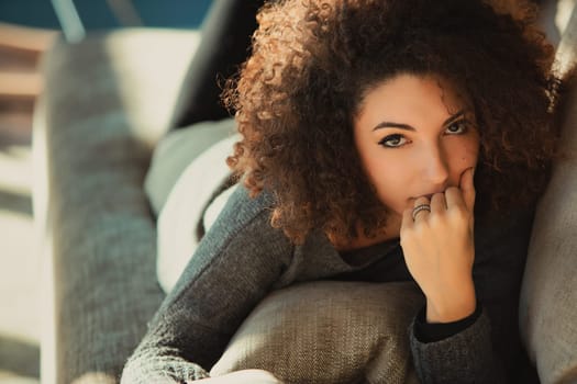 Young woman with curly hair relaxes on a cozy sofa in her living room, lost in thought and enjoying a peaceful moment of tranquility