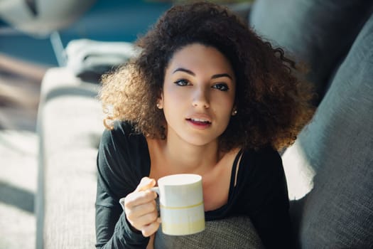 Young woman with curly hair is taking a break from her day, enjoying a cup of coffee while relaxing on a comfortable couch