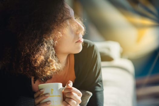 Young woman enjoying a coffee break at home on her comfy couch, gazing out the window in peaceful morning sunlight