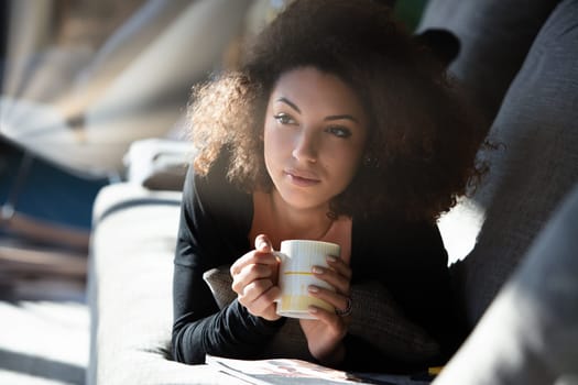 Young woman relaxes at home, sipping coffee and reading a magazine on her cozy sofa, enjoying a peaceful morning flooded with natural light