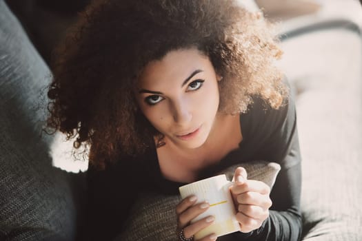 Young woman with curly hair enjoys a cozy moment at home, savoring a cup of coffee on her sofa. The natural light creates a tranquil atmosphere, perfect for self care and relaxation