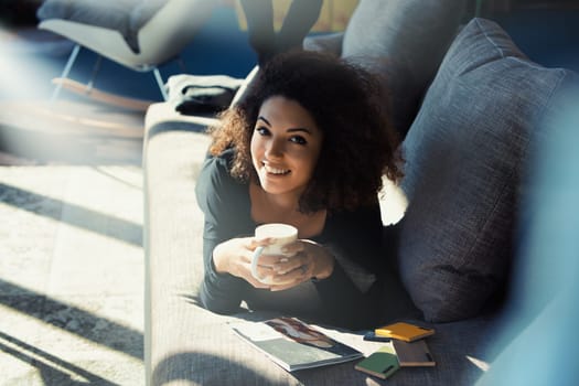 Young woman enjoys coffee on her couch, working from home in a sunny living room, taking a peaceful break in her cozy apartment