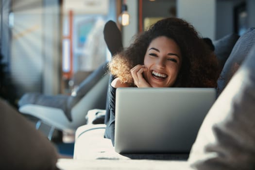 Young woman with curly hair is laughing while using a laptop on the couch, enjoying the comforts of working from home