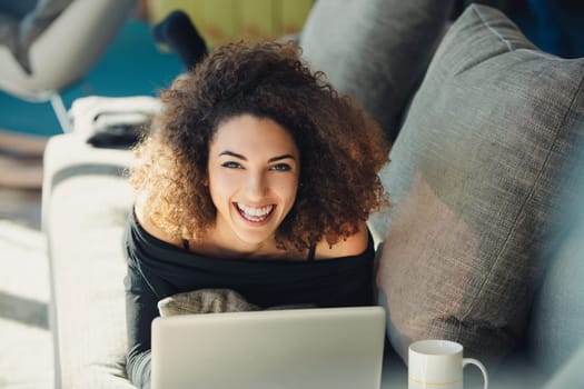 Young woman with curly hair is lying on her stomach on a couch and using a laptop, she is smiling and appears to be enjoying herself