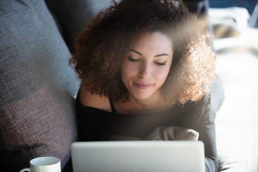 Young woman with curly hair works on a laptop from home, lying comfortably on a couch in a cozy living room. She looks relaxed and focused, enjoying the modern lifestyle of remote work