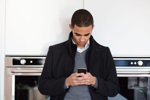 Young businessman is standing in a modern kitchen, wearing a black coat and holding his smartphone. He is looking at the screen of the phone with a serious expression