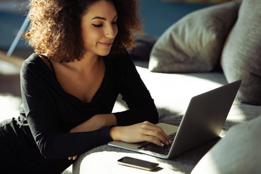 Young woman is lying comfortably on her sofa, working on her laptop in her living room. She is smiling and appears to be enjoying her work