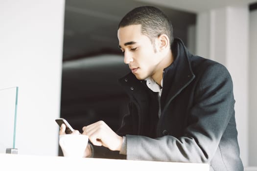 Young man sits at a table in a modern home, using his smartphone during a coffee break, looking focused and serious