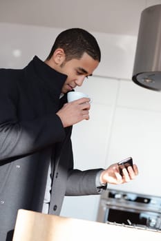 Young businessman enjoys coffee and checks his phone in a modern kitchen, balancing work and leisure effortlessly at home