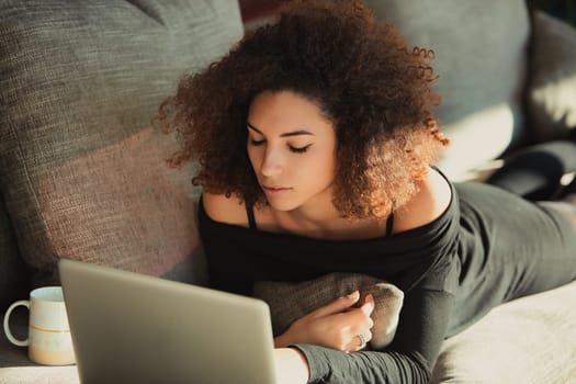 Young woman is lying on her couch working on a laptop with a cup of coffee nearby. She is enjoying the comforts of home while getting some work done