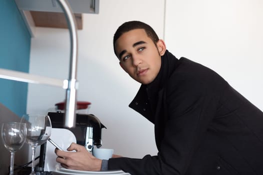 In a modern kitchen, a young entrepreneur washes dishes, lost in thought. Water flows as he scrubs clean, reflecting daily responsibility. Image shows diverse domestic life