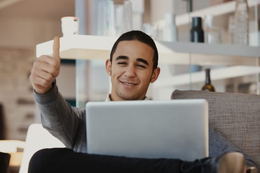 Young entrepreneur is relaxing on his couch at home, working on his laptop and giving a thumbs up, happy with his success