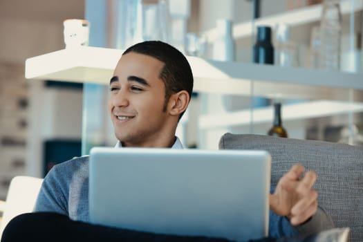 Young man is sitting on a sofa in his living room, working from home and having a video call on his laptop. He is smiling and gesturing with his hand