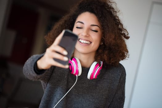 Young woman in a cozy kitchen takes a happy selfie with pink headphones, enjoying music and sharing her joy on social media