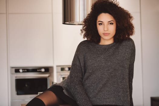 Young woman with curly hair sits comfortably in a bright modern kitchen, exuding confidence and relaxation with a cheerful smile