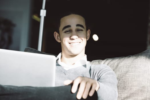 Young man happily smiles while using his laptop on a sofa at home, enjoying a break and connecting online in a cozy living room with natural light