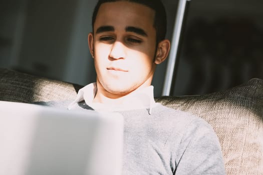 Young man is working at his laptop while sitting on a sofa at home, bathed in warm sunlight streaming through a window