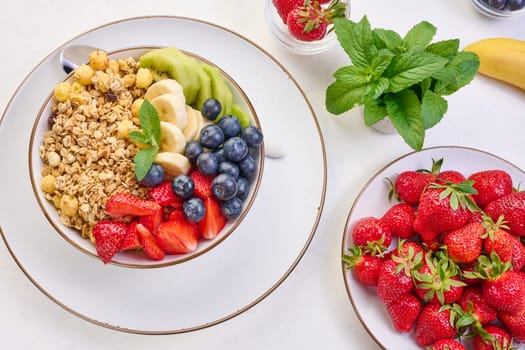 Granola with strawberries, kiwi, banana and blueberries in a round plate on a white table. Healthy and tasty food, top view