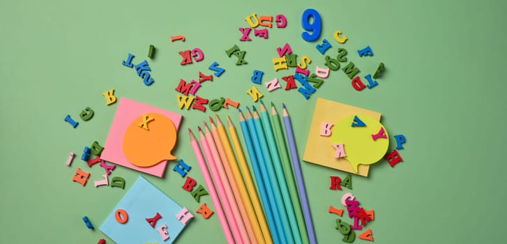 Stationery laid out on a green background. Multi-colored pencils ,  sticky notes and wooden letters, preparing for study or work, organizing workspace