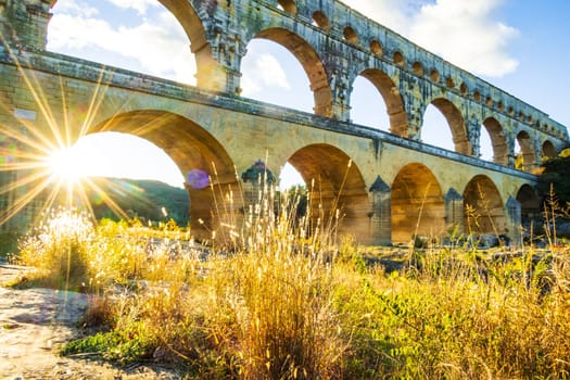 The magnificent Pont du Gard backlit by the setting sun. Ancient Roman three-tiered aqueduct bridge built in the 1st century AD to carry water to the Roman colony of Nemausus (Nîmes). Photograph taken in Vers-Pont-du-Gard, Provence, southern France.