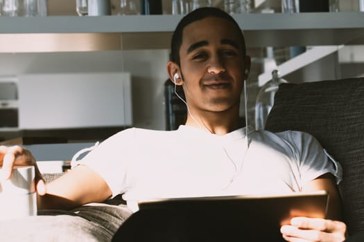 Young man is spending his free time at home, listening to music, drinking coffee and browsing the internet on his tablet