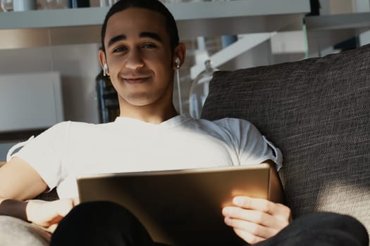 Young man is sitting on a comfortable sofa in his living room, enjoying some downtime with his digital tablet and listening to music through his earbuds