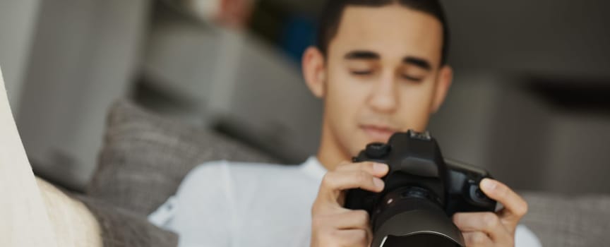 Young man adjusts a professional camera on a sofa at home, immersed in photography. Natural light enhances his creative focus
