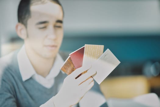 Seated young man with short hair studies color samples, wearing a gray sweater over a white shirt. Sunlight streams in, casting shadows on his focused face and brightening the modern space
