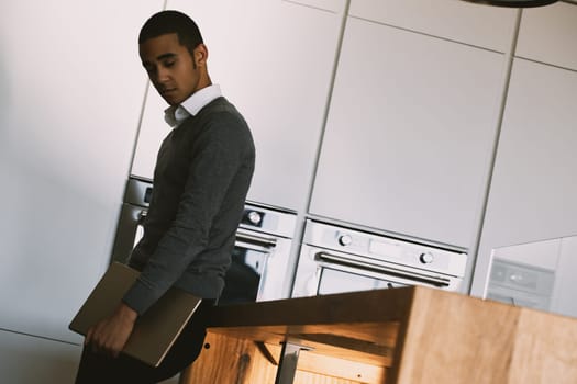 Young man in a gray sweater and white shirt holds a laptop with a confident expression. He stands in a modern kitchen, feeling cool and composed. The stylish environment enhances his demeanor