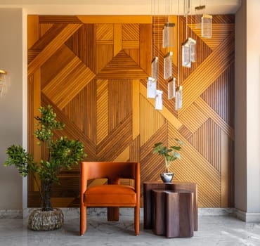 Simple and elegant shot of a modern style orange chair and coffe table in front of a decorated wood cladding wall, with hanging lights and a potted green plants