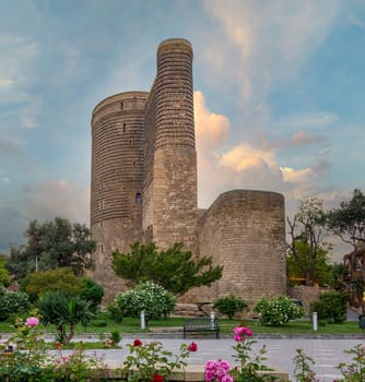 The Maidan Tower in Baku, Azerbaijan stands tall against a backdrop of clouds in the evening sky. Lush greenery surrounds the historic structure.