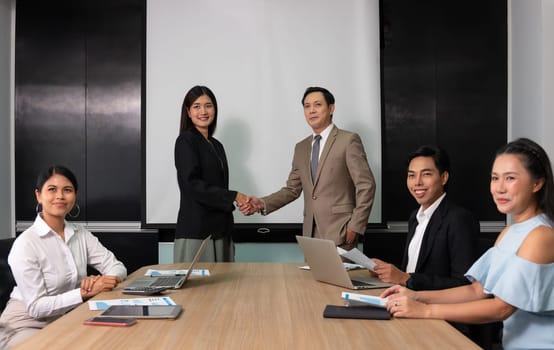 Asian happy successful two businessman and businesswoman partnership hands shaking and greeting together while standing in board room with group of businesspeople sitting on desk. Business agreement achievement partnership deal