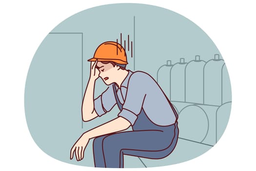 Tired man working in factory sits near production equipment holding head because of nervous job. Nervous factory worker in safety helmet needs psychological support due to poor working conditions