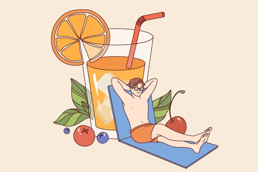 Man relaxed on beach and drinks orange juice from giant glass, enjoying sunny weather. Happy guy sunbathing near pool and enjoy life traveling to resorts with cool hotels for tourists