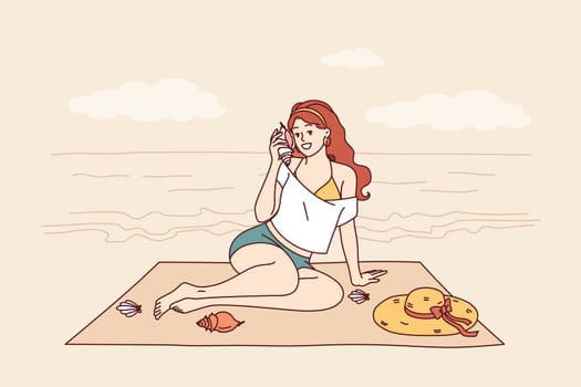 Woman sits on beach and listens to sound of sea from shell, enjoying summer vacation at tropical resort. Tourist girl sunbathes on beach of paradise island in ocean during weekend.