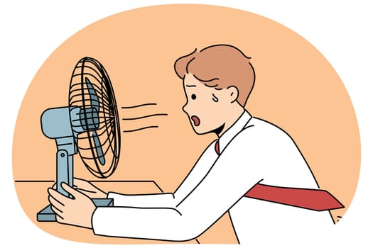 Sweaty man sits in front of fan enjoying cold wind from electric climate control equipment. Young businessman with fan suffers from lack of air conditioning and ventilation in office