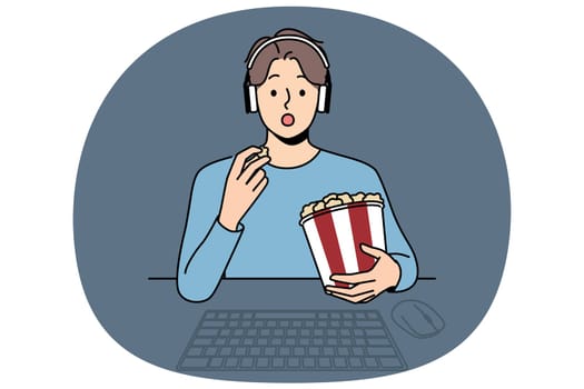 Man with package of popcorn watches movie sitting at table with computer and looks at screen intrigued. Funny guy in headphones is relaxing watching new movie blockbuster or popular tv series