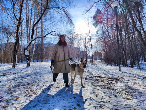 A girl in winter attire walks her German Shepherd dog through a snowy park, surrounded by trees, enjoying a sunny day.
