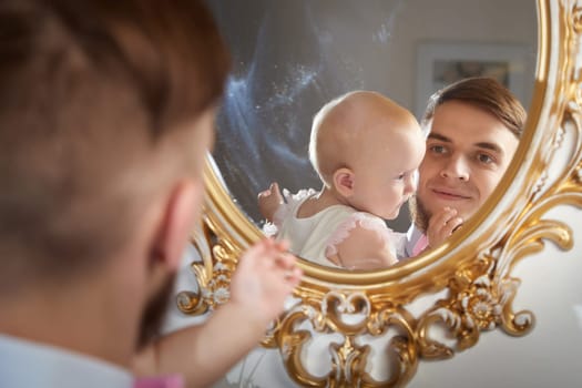 A father smiles while holding his baby daughter, who plays with her fingers, in a warm, inviting space.